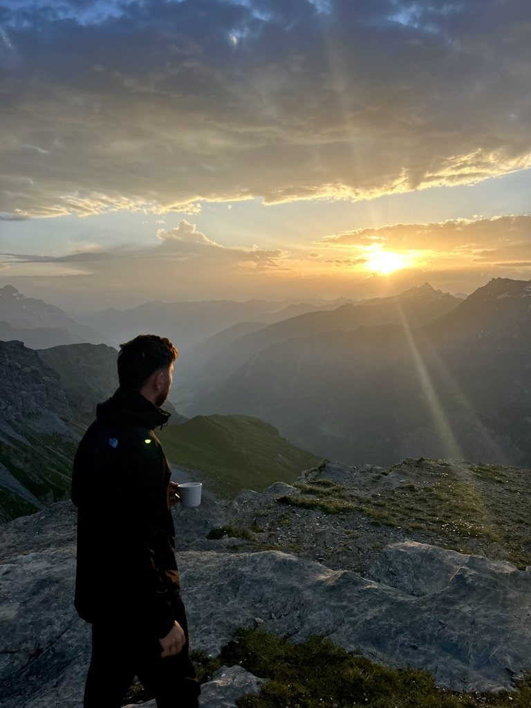 Julian steht mit einer Tasse in der Hand auf einem Felsgrat und schaut in den Sonnenaufgang über den Bergen