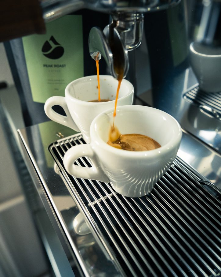 Espresso läuft in zwei weisse Tassen, dahinter die PEAK ROAST Packung